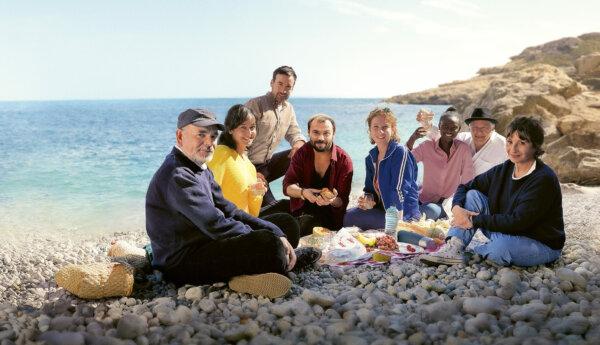 en stillbild ur en film där en åttapersonersfamilj sitter på en stenig strand