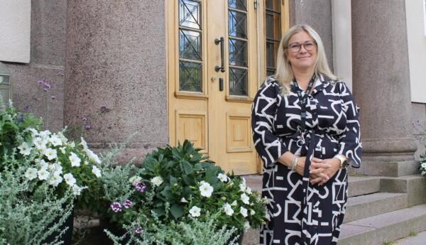 En kvinna står framför stadshuset i Ekenäs. Hon är stadens ny undervisningschef. Bredvid henne finns tre stycken större blomkrukor med blommor i.