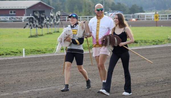 Två kvinnor och en man med käpphästar promenerar på travbana